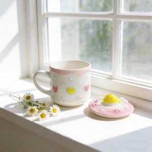 Peut inclure: Une tasse en c&eacute;ramique blanche avec un bord rose et des motifs en forme de c&oelig;ur roses et jaunes. La tasse a un couvercle assorti avec un poussin jaune. Des marguerites sont plac&eacute;es pr&egrave;s de la tasse sur un rebord de fen&ecirc;tre.