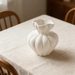 Könnte beinhalten: Eine weiße, strukturierte Keramikvase mit einer einzigartigen, bauchigen Form und einem gerüschten Rand. Die Vase steht auf einer neutralfarbenen Tischdecke, im Hintergrund Holzstühle. Die Vase hat eine strukturierte Oberfläche.