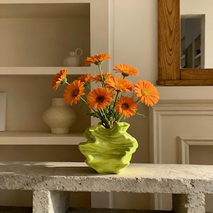 Könnte beinhalten: Ein lebendiges Arrangement aus orangefarbenen Gerbera-Gänseblümchen in einer einzigartigen, limettengrünen Keramikvase mit gewelltem Rand. Die Vase steht auf einer Betonoberfläche, vor einem neutralen Hintergrund mit Regalen und einem dekorativen Spiegel.