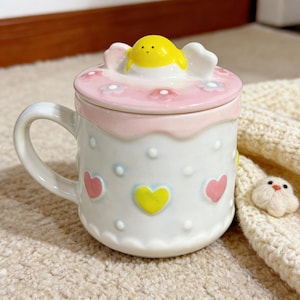Peut inclure: Une tasse en c&eacute;ramique blanche avec un couvercle, d&eacute;cor&eacute;e de c&oelig;urs et de fleurs. Le couvercle est rose avec une figurine de poussin jaune sur le dessus. La tasse a une anse et est pos&eacute;e sur une surface beige.
