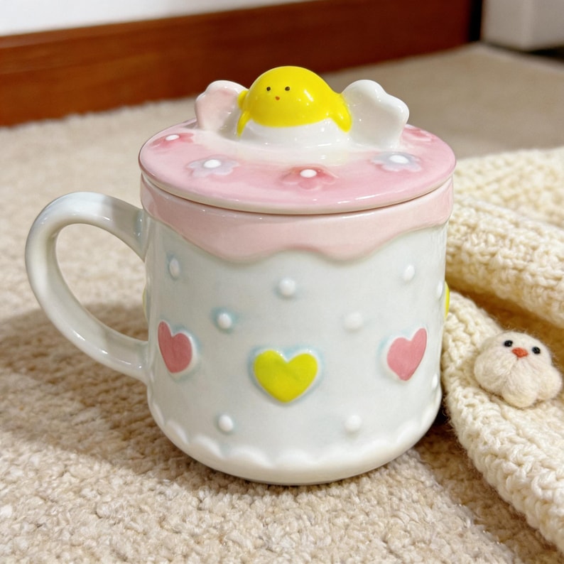 Peut inclure: Une tasse en c&eacute;ramique blanche avec un couvercle, d&eacute;cor&eacute;e de c&oelig;urs et de fleurs. Le couvercle est rose avec une figurine de poussin jaune sur le dessus. La tasse a une anse et est pos&eacute;e sur une surface beige.