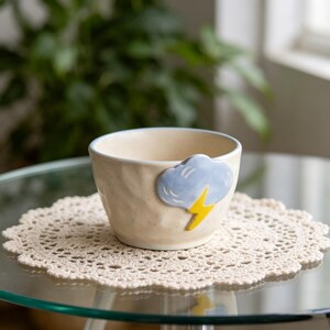 May include: A cream-colored ceramic mug with a blue cloud and yellow lightning bolt design. The mug sits on a beige crocheted doily on a glass table. The mug has a textured surface.