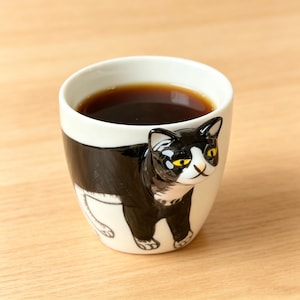 May include: A white ceramic mug with a black and white cat design. The mug is filled with dark liquid, likely coffee or tea. The cat's face and body wrap around the mug, with yellow eyes and a playful expression. The mug sits on a light brown wooden surface.