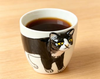 Peut inclure: Une tasse en céramique blanche avec un motif de chat noir et blanc. La tasse est remplie d'un liquide foncé, probablement du café ou du thé. Le visage et le corps du chat sont représentés sur la tasse, avec des yeux jaunes. La tasse est posée sur une surface en bois brun clair.