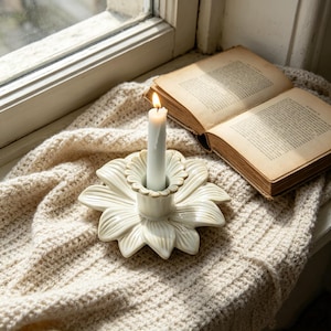 May include: A lit white candle in a cream-coloured, flower-shaped candle holder. An open, aged book rests beside the candle on a cream-coloured knitted blanket, near a window. The candle holder is ceramic.
