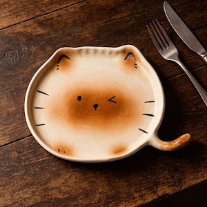 May include: A cat-shaped ceramic plate with a brown face and cream-coloured edges. The plate has black whiskers, ears, and a winking eye. A tail extends from the side. A fork and knife are next to the plate.