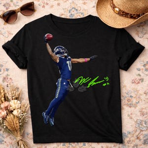 Puede incluir: Camiseta negra con un jugador de fútbol americano con uniforme azul atrapando un balón. El jugador lleva un casco con el número 11. La camiseta tiene una firma verde y un diseño de hojas.