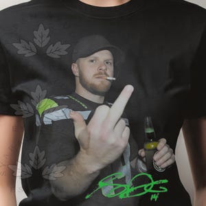 May include: Black t-shirt featuring a photo of a man wearing a hat, smoking a cigarette, and holding a beer bottle. The man is giving the middle finger. Green signature and the number 14 are visible.