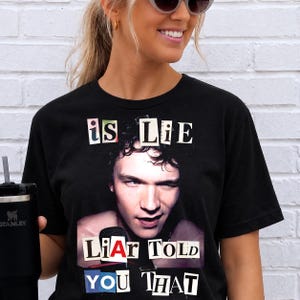 Könnte beinhalten: Schwarzes T-Shirt mit einem Grafik eines Mannes, der zwinkert, und den Worten "is Lie" über seinem Kopf. Unter dem Bild steht der Text "Liar Told You That".