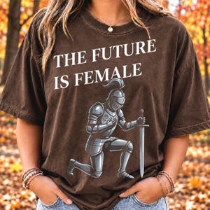 Op de afbeelding: Een donkerbruin T-shirt met korte mouwen en de witte tekst THE FUTURE IS FEMALE boven een afbeelding van een ridder in zilveren harnas die knielt en een zwaard vasthoudt. De ridder heeft een klein rozenembleem op het borstpantser.