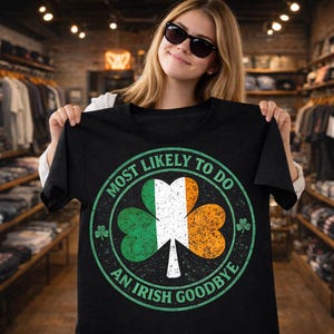Könnte beinhalten: Schwarzes T-Shirt mit einem Kleeblatt-Design in Grün, Weiß und Orange. Der Text "MOST LIKELY TO DO AN IRISH GOODBYE" umrundet das Kleeblatt. Das T-Shirt wird von einer Person präsentiert.