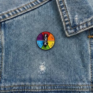 May include: A round enamel pin with a colorful design and the words "FUCK OFF" in the center. The pin is divided into sections with phrases like "YOU", "ME", and "THIS". The pin is attached to a denim jacket.