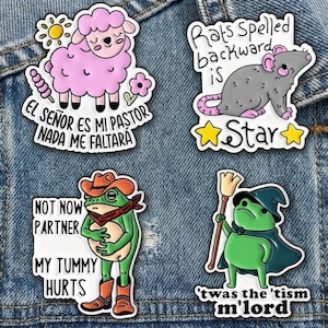 May include: Four colorful enamel pins on a denim jacket. The pins feature a pink sheep with text, a gray rat with text, a green frog in cowboy attire with text, and a green frog in a cape with a broom and text.