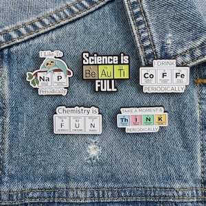 May include: A collection of enamel pins with science-themed designs. The pins feature periodic table elements and phrases like "Science is FULL" and "Chemistry is FUN." The pins are attached to a denim jacket.
