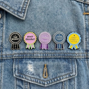 Puede incluir: Cinco pines de esmalte con frases humorísticas, incluyendo "The Worst", "Overthinker" y "Only Cried a Little". Los pines son de varios colores, como negro, rosa, morado y azul, y están sujetos a una chaqueta vaquera.