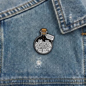 Könnte beinhalten: Runder Emaille-Pin in Schwarz und Weiß, der eine Flasche mit Katzen-Gesichtern darstellt. Die Flasche hat einen Korken und ein Etikett mit der Aufschrift "Happiness Potion". Der Pin ist auf einer blauen Jeansjacke angebracht.