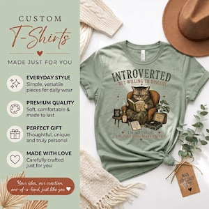 May include: A sage green t-shirt featuring a graphic of a cat sitting in an armchair reading a book. The text reads: INTROVERTED BUT WILLING TO DISCUSS and I'M NOT RUDE: I'M JUST CONSERVING ENERGY. The design includes a small table, a plant, and a record player.
