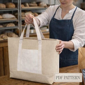 May include: A beige tote bag with a white striped pocket and matching handles. The bag is held by a person in a bakery setting. The text "PDF PATTERN" is visible on a tag.