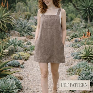 Op de afbeelding: Een bruine linnen tuinbroekjurk met een vierkante halslijn, grote voorzak en zijzakken. De jurk wordt over een witte top gedragen. De afbeelding bevat de tekst "PDF PATTERN". De jurk is knielang.