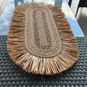 Peut inclure: Nappe ovale en fibres naturelles avec un centre tissé et des bords frangés. Le napperon est de couleur beige et marron et est placé sur une table en verre. Le napperon est conçu pour un usage décoratif.