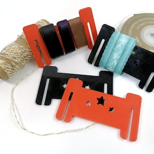 Puede incluir: Un surtido de materiales para manualidades que incluyen carretes de hilo, cintas y soportes para cintas naranjas y negras con recortes de estrellas. También se ve un rollo de cinta. Los artículos están dispuestos sobre una superficie blanca.