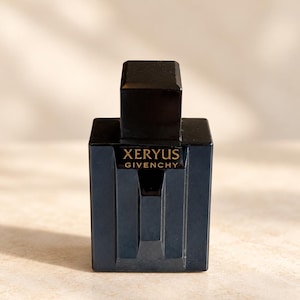 Könnte beinhalten: Ein rechteckiger, dunkelblauer Glasparfümflakon mit passendem schwarzem Verschluss. Auf der Vorderseite des Flakons steht der Schriftzug XERYUS GIVENCHY in goldenen Buchstaben. Der Flakon hat ein strukturiertes, geometrisches Design.