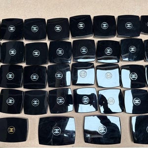 Op de afbeelding: Een verzameling zwarte vierkante compactpoeders met een wit logo in het midden. De compactpoeders zijn gerangschikt in rijen en kolommen op een lichtbruin oppervlak. Het logo is een gestileerde dubbele C.