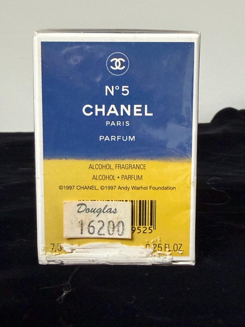 May include: A rectangular perfume box with a blue and yellow design. The box features the Chanel logo and the text "N&deg;5 CHANEL PARIS PARFUM." The box also includes the text "ALCOHOL FRAGRANCE ALCOHOL PARFUM" and "25 FL.OZ."