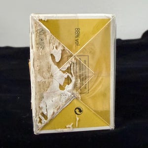 May include: A rectangular, gold-colored box with a partially torn, peeling label. The box has a geometric design on the top and the text "88% vol." is visible. The box is set against a dark background.