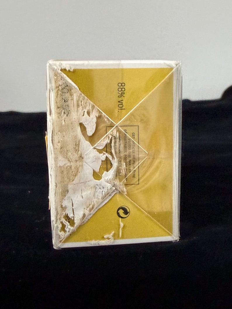May include: A rectangular, gold-colored box with a partially torn, peeling label. The box has a geometric design on the top and the text "88% vol." is visible. The box is set against a dark background.