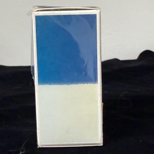 May include: A rectangular box with a two-tone design. The top half is a solid blue, and the bottom half is a light cream color. The box is wrapped in clear plastic.