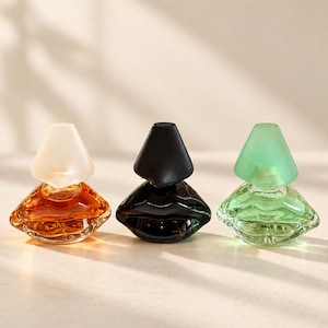 Op de afbeelding: Drie glazen parfumflesjes in de vorm van lippen, elk met een dop in een eigen kleur. Het linkerflesje is amberkleurig met een witte matte dop, het middelste is zwart met een zwarte dop en het rechterflesje is lichtgroen met een bijpassende groene dop.