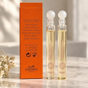 Può includere: Due flaconi di profumo in vetro, alti e sottili, con tappi sferici bianchi, riempiti di liquido giallo chiaro. Accanto si trova una scatola rettangolare arancione con l'etichetta Hermès Paris, che elenca profumi come Rose Ikebana e Osmanthe Yunnan, 2 x 4 ml.