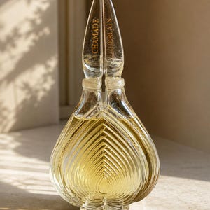 Könnte beinhalten: Ein transparenter Glasparfümflakon mit herzförmigem Körper, vertikalen Rillen und einem hohen, spitzen Verschluss. Der Flakon enthält eine goldene Flüssigkeit und trägt die Aufschrift CHAMADE und GUERLAIN in goldenen Buchstaben auf dem Glas.