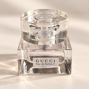 Puede incluir: Un frasco de perfume de vidrio transparente con base cuadrada y tapa cilíndrica. La etiqueta frontal presenta el texto GUCCI Eau de Parfum II en letras negras. El frasco contiene un líquido rosa claro y tiene un diseño transparente y reflectante.