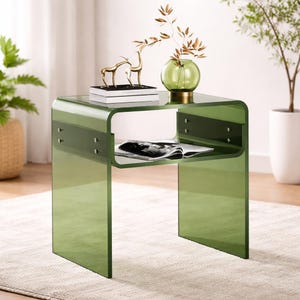 May include: A modern, green acrylic side table with a curved design. The table features a lower shelf and is adorned with decorative items, including a gold deer figurine and a green glass vase with dried branches. The table is placed on a light-colored rug.