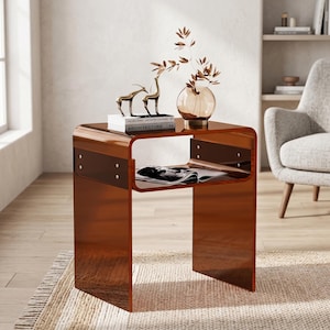 May include: A translucent amber-colored acrylic side table with a curved C-shaped design. The table features a horizontal shelf supported by metal hardware on the sides, providing an open storage space for books and decorative items.