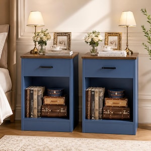 May include: Two matching blue nightstands with a dark wood top, each featuring a single drawer with a black handle and an open storage shelf below. The shelves contain stacked books and small vintage-style suitcases. Each nightstand holds a lamp.