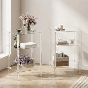 May include: Two clear acrylic side tables with three shelves each. The tables are filled with decorative items, including books, candles, and flowers. The tables are placed on a light-colored wooden floor.