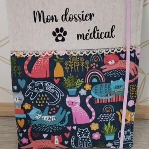 May include: A fabric cover with the text "Mon dossier médical" and a paw print. The cover features a colourful cat pattern with pink, blue, and orange cats on a dark blue background. A pink elastic band secures the cover.