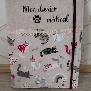 May include: A beige fabric medical file holder with a cat pattern. The holder has a lace trim and a burgundy elastic closure. The text "Mon dossier médical" and a paw print are printed on the top.