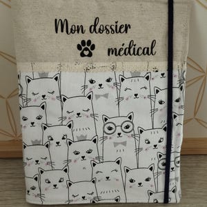 May include: A fabric document holder featuring a cat design. The top section is beige with the text "Mon dossier médical" and a paw print. The lower section has a repeating pattern of white cats with black outlines and pink accents. A navy blue elastic band secures the holder.