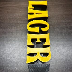 以下が含まれることがあります： 黄色い文字で「LAGER」と書かれた3Dプリントのサイン。文字は縦に積み重ねられ、下部に黒いベースがあります。バーやホームバーに最適で、プラスチックなどの素材で作られている可能性があります。