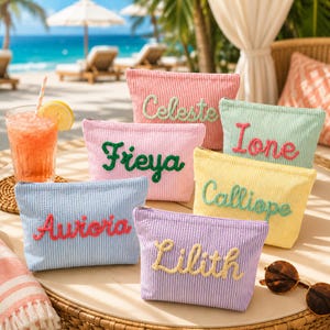 May include: A collection of colorful, striped cosmetic bags, each embroidered with a different name. The bags are in shades of pink, blue, yellow, green, and purple. A refreshing drink with a straw and sunglasses are also visible, suggesting a beach or vacation theme.