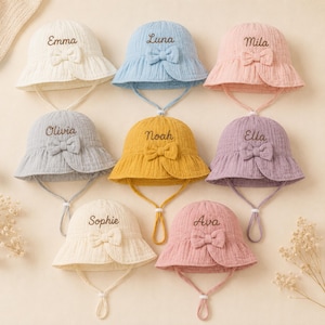 Puede incluir: Una colección de ocho sombreros de sol para bebés en varios colores pastel, cada uno con un lazo y un nombre bordado en la parte delantera. Los sombreros tienen correas para la barbilla y un ala con volantes.