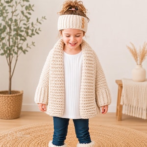 May include: A young child wearing a cream-colored, chunky knit cardigan and matching headband with wooden buttons. The child is also wearing a white shirt, blue jeans, and white fuzzy boots. The outfit is handmade and suitable for cold weather.