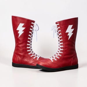 May include: A pair of mid-calf lace-up boots in red leather featuring a white lightning bolt graphic on the outer side. The boots have white laces, a rounded toe, and a flat black sole, providing a structured look with a high-top design.