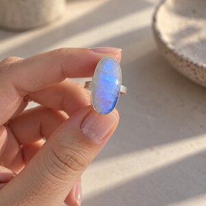 May include: A silver ring featuring a large, oval-shaped moonstone cabochon. The gemstone displays a translucent, milky white appearance with a distinct, vibrant blue adularescence or flash across its surface. The stone is set in a simple, polished silver bezel mount with a plain, slender silver band.