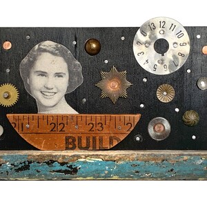 May include: A black wooden plaque with a vintage photo of a woman, a ruler with the word "BUILD" printed on it, and various metal and plastic embellishments. The plaque is mounted on a weathered wooden base.