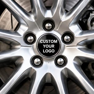 May include: A silver multi-spoke car wheel rim featuring a central circular cap with the black text CUSTOM YOUR LOGO. The design includes five lug nuts arranged in a circle around the centre, with a visible brake disc and caliper behind the spokes.